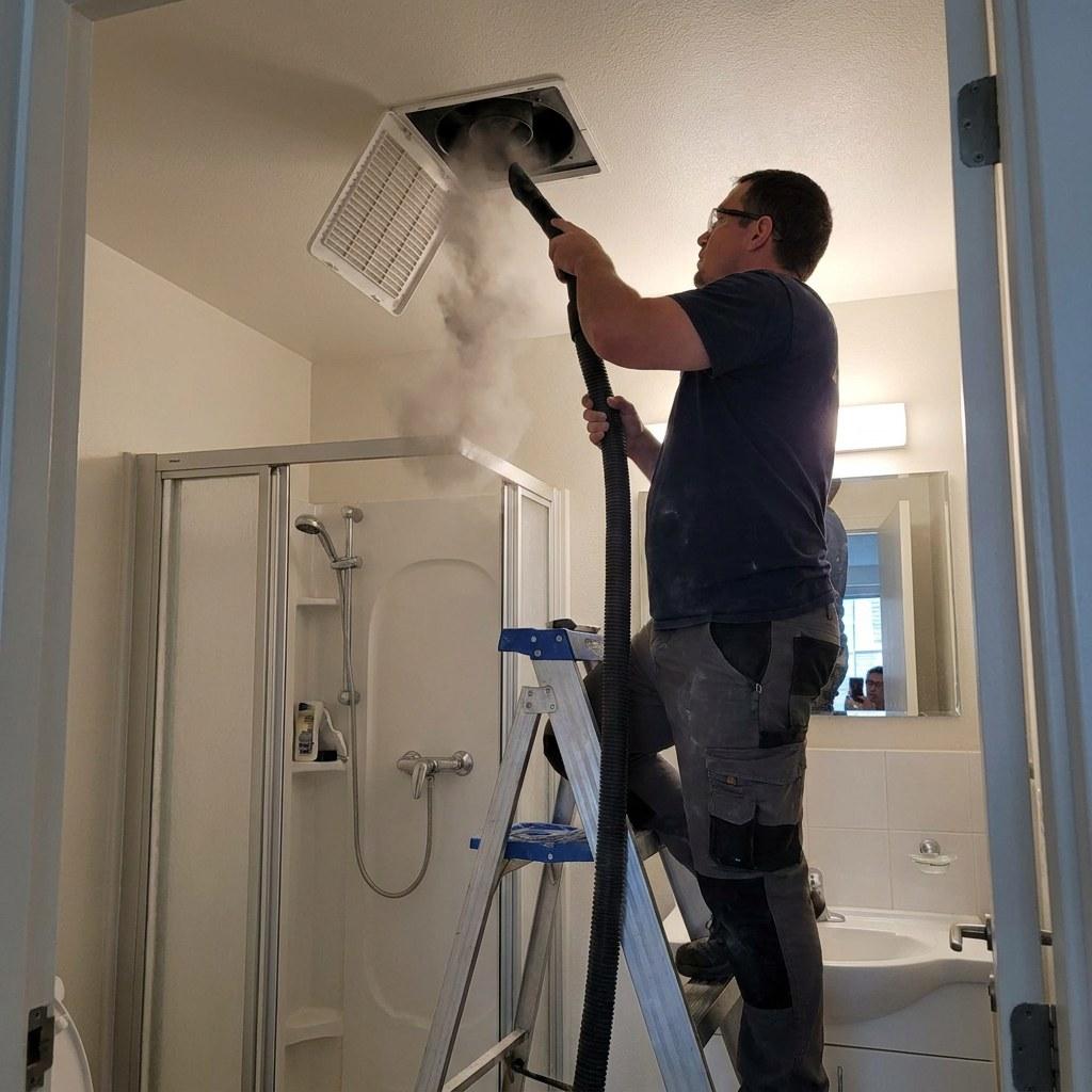 A person standing on a step stool removing the plastic cover from a bathroom ceiling exhaust fan for cleaning
